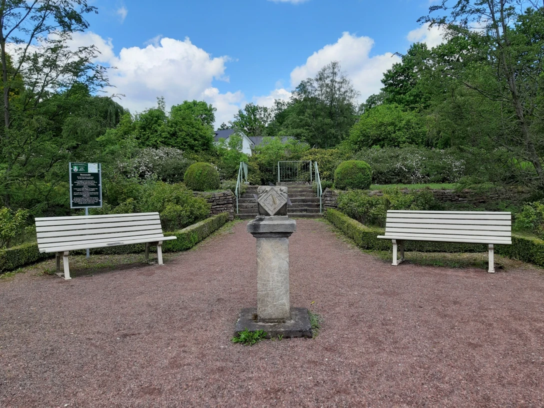 Steinerne Sonnenuhr auf einem Sockel, flankiert von weißen Gartenbänken in einer gepflegten Parkanlage.