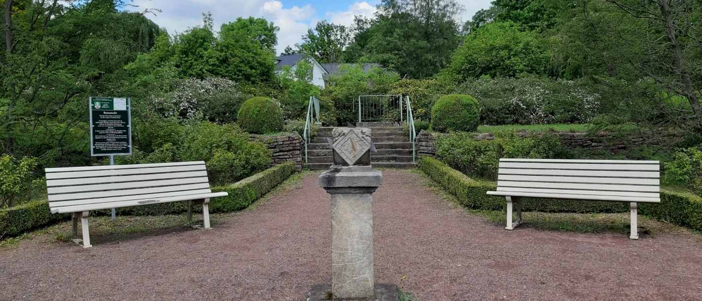 Steinerne Sonnenuhr auf einem Sockel, flankiert von weißen Gartenbänken in einer gepflegten Parkanlage.