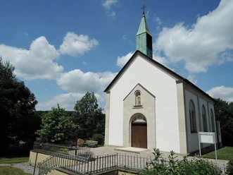 St. Josef Kapelle in Feldrom mit weißer Fassade, grünem Turm und umgeben von üppigem Grün.