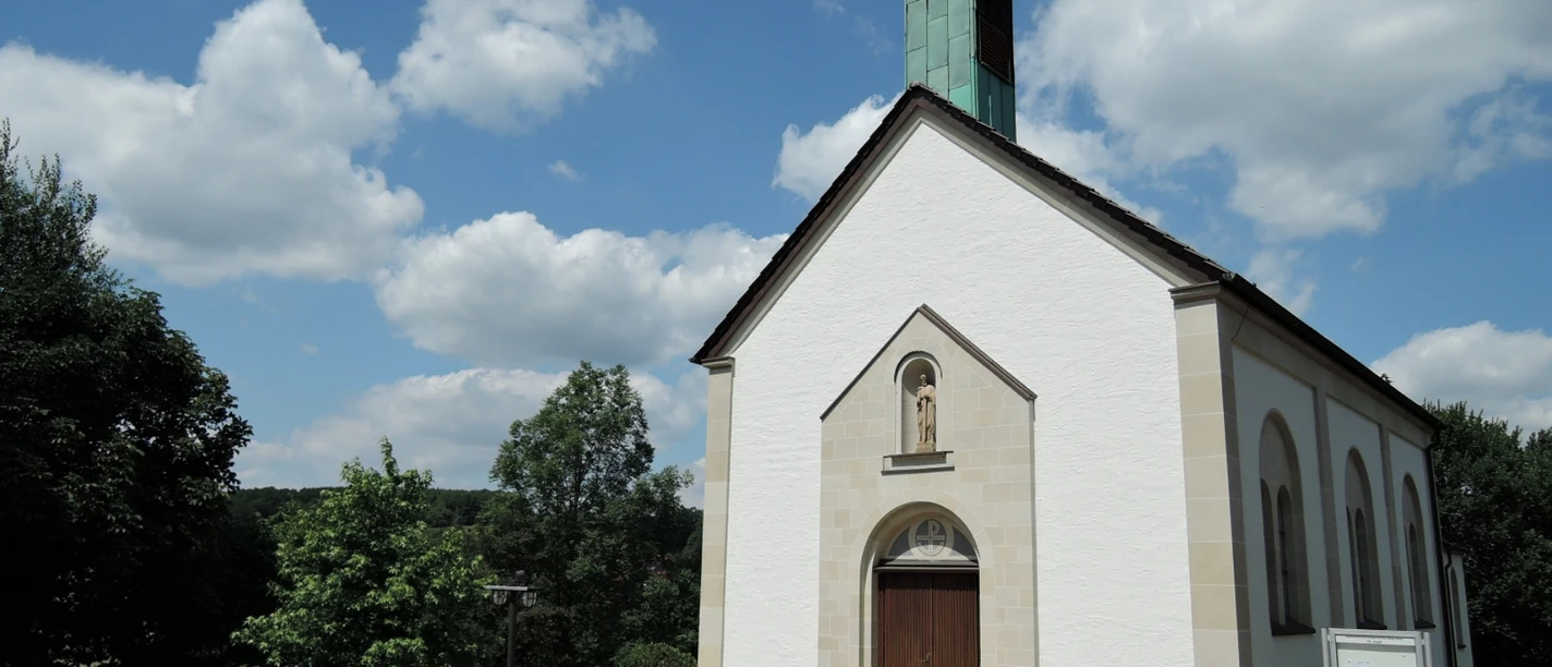 St. Josef Kapelle in Feldrom mit weißer Fassade, grünem Turm und umgeben von üppigem Grün.
