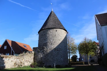 Eulenturm Runder, steinerner Turm mit spitzem Dach, umgeben von Mauerresten und modernen Gebäuden im Hintergrund.