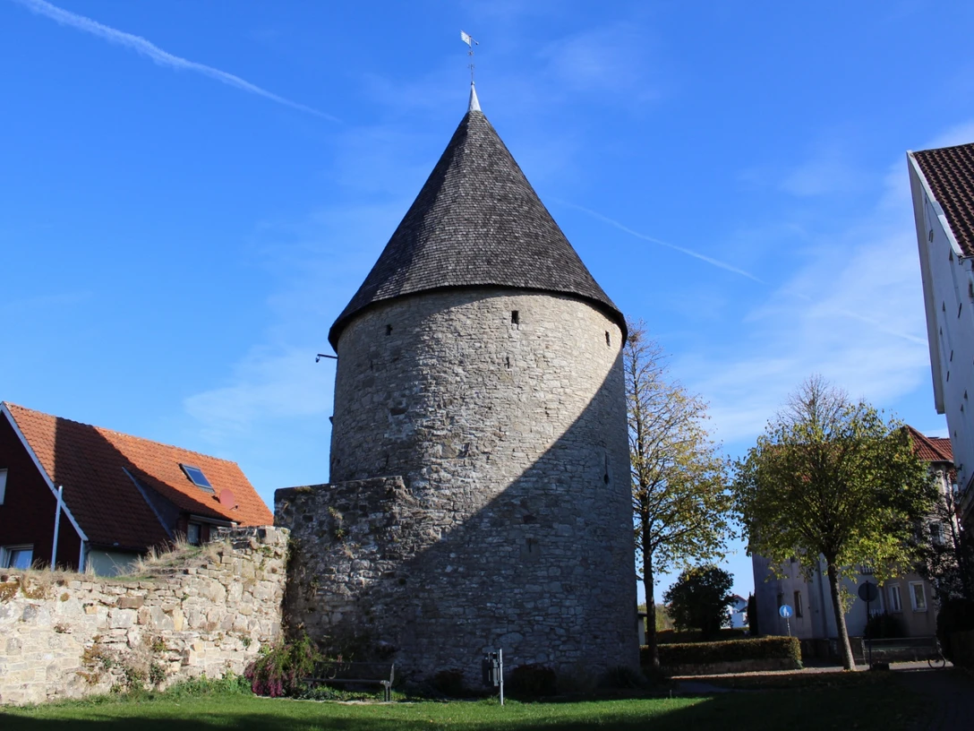 Eulenturm Runder, steinerner Turm mit spitzem Dach, umgeben von Mauerresten und modernen Gebäuden im Hintergrund.