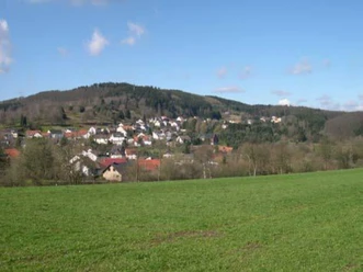 Grüne Wiesen führen zu einem kleinen Dorf vor bewaldeten Hügeln unter einem blauen Himmel.