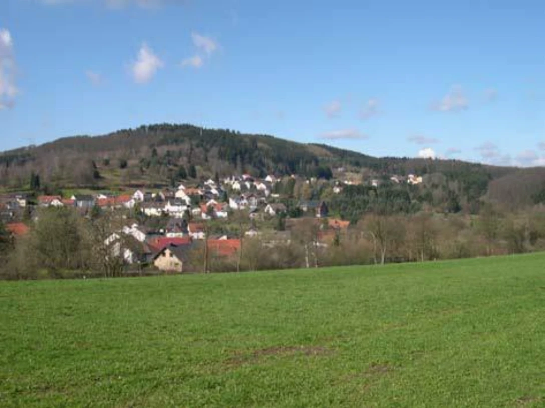 Stemberg Grüne Wiesen führen zu einem kleinen Dorf vor bewaldeten Hügeln unter einem blauen Himmel.