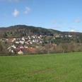 Stemberg Grüne Wiesen führen zu einem kleinen Dorf vor bewaldeten Hügeln unter einem blauen Himmel.