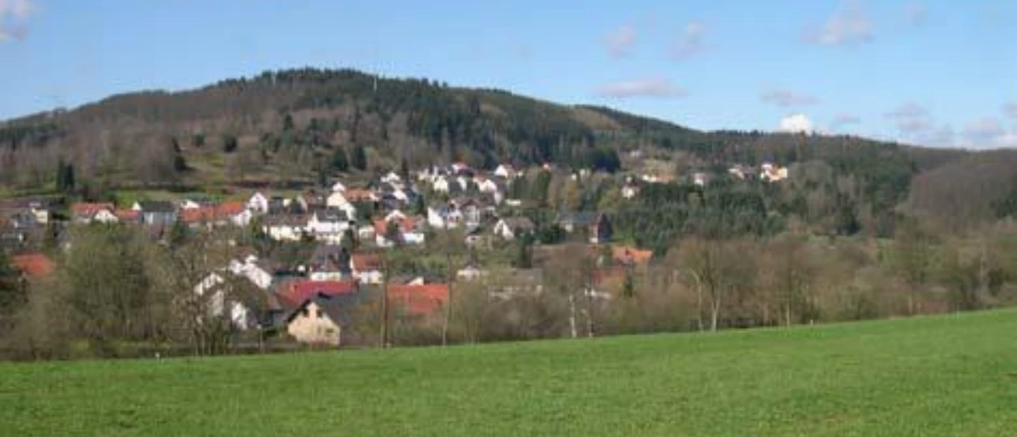 Stemberg Grüne Wiesen führen zu einem kleinen Dorf vor bewaldeten Hügeln unter einem blauen Himmel.