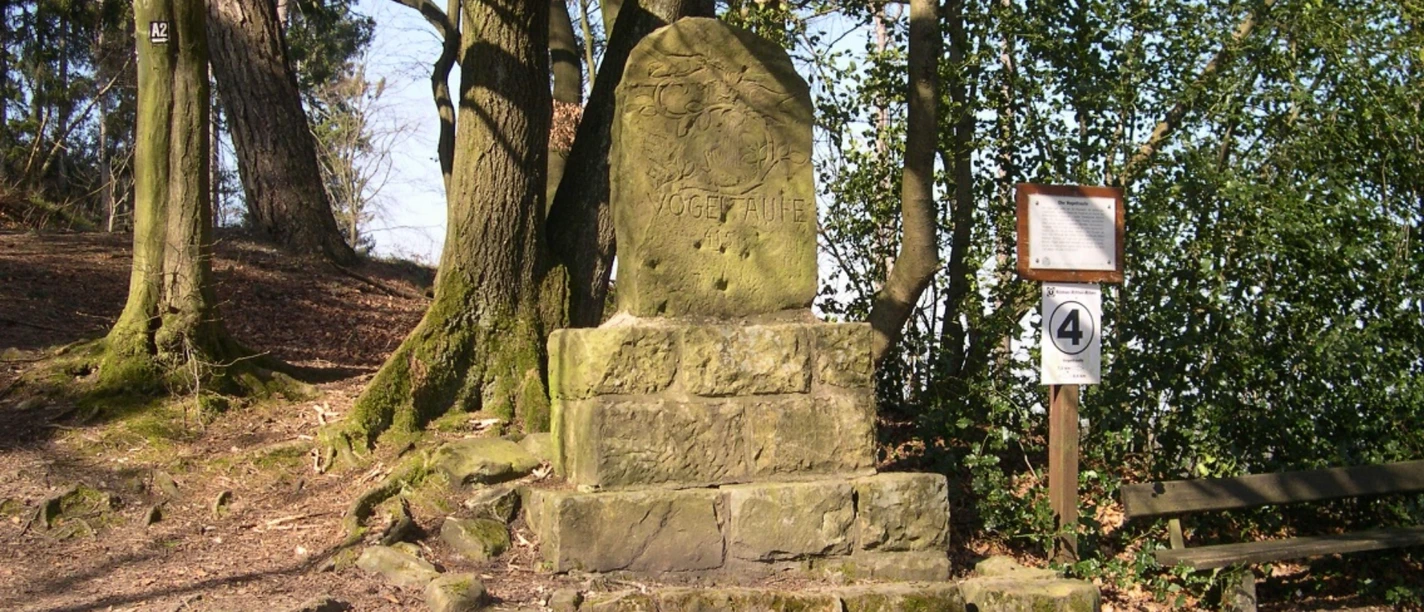 Steinmonument umgeben von Bäumen mit einer Tafel und Wegweiser, Standort Vogeltaufe.