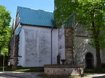 Pfeifenkumpbrunnen am Rand einer alten Kirche, umgeben von Bäumen, mit blauem Himmel im Hintergrund.