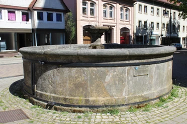 Ein runder, steinerner Brunnen mit verwitterter Oberfläche, umgeben von historischen Gebäuden.