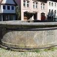 Pfeifenkump Ein runder, steinerner Brunnen mit verwitterter Oberfläche, umgeben von historischen Gebäuden.