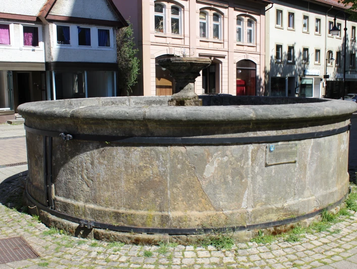 Pfeifenkump Ein runder, steinerner Brunnen mit verwitterter Oberfläche, umgeben von historischen Gebäuden.