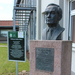 Büste von Wilhelm Rebbe aus Bronze auf grauem Sockel vor einer modernen Gebäudefassade.