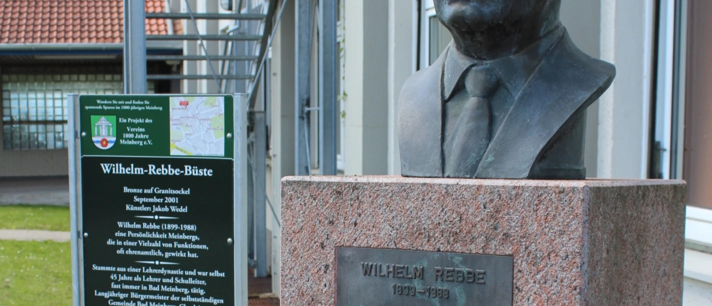 Wilhelm-Rebbe-Büste Büste von Wilhelm Rebbe aus Bronze auf grauem Sockel vor einer modernen Gebäudefassade.