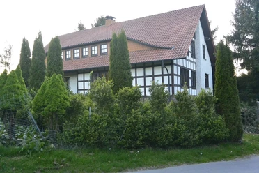 wiehen-inn-1.jpg Fachwerkhaus mit rotem Ziegeldach, umgeben von grünen Bäumen und Hecken an einer ruhigen Straße.