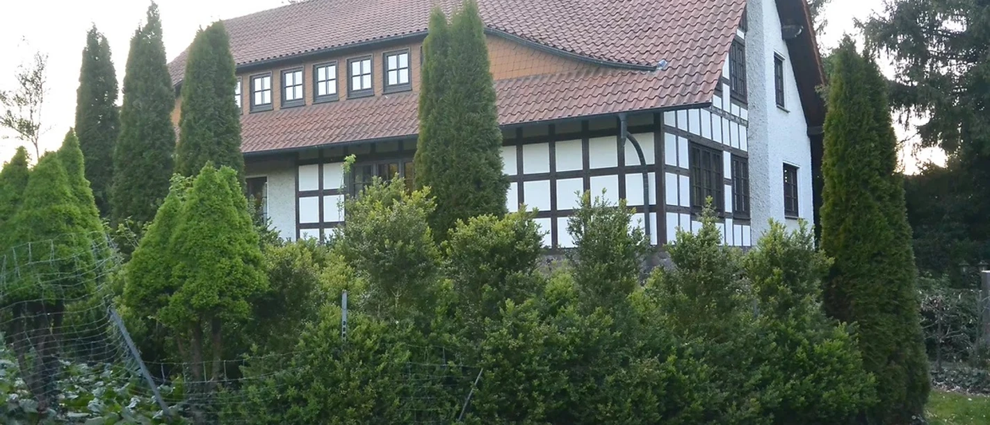 wiehen-inn-1.jpg Fachwerkhaus mit rotem Ziegeldach, umgeben von grünen Bäumen und Hecken an einer ruhigen Straße.