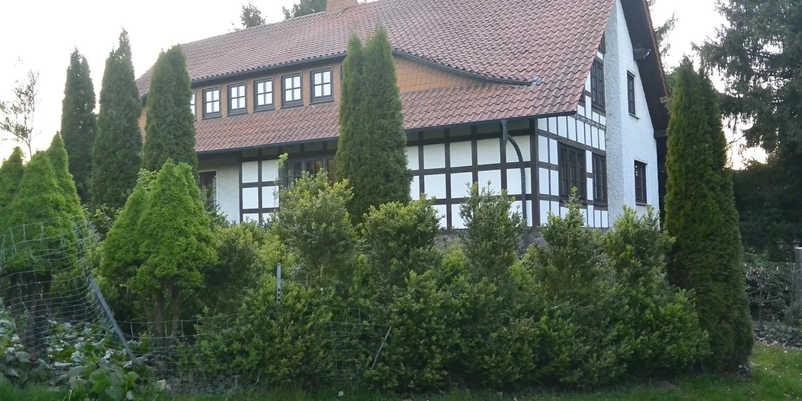 wiehen-inn-1.jpg Fachwerkhaus mit rotem Ziegeldach, umgeben von grünen Bäumen und Hecken an einer ruhigen Straße.