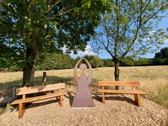 Metallengel mit erhobenen Armen zwischen zwei Holzbänken auf Holzschnitzel, umgeben von Bäumen.