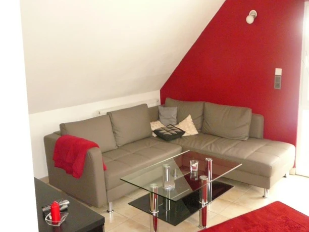 Rahe-1.jpg Modernes Wohnzimmer mit grauem Ecksofa, Glastisch und roten Akzenten unter einer Dachschräge.