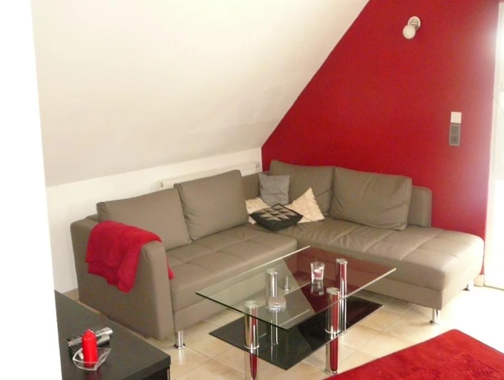 Rahe-1.jpg Modernes Wohnzimmer mit grauem Ecksofa, Glastisch und roten Akzenten unter einer Dachschräge.