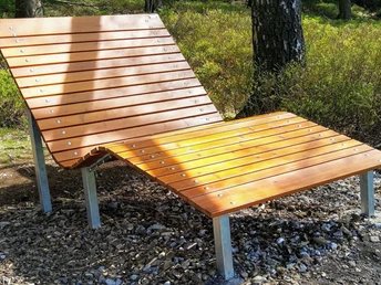 Hölzerne Liege auf Schotteruntergrund im Wald, beleuchtet von Sonnenstrahlen, umgeben von Bäumen.