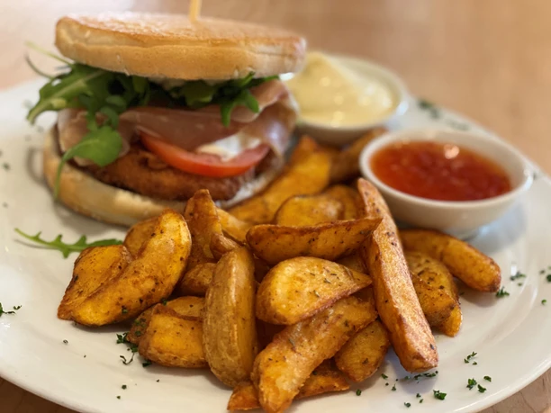 Ein saftiger Burger mit Prosciutto und Gemüse, serviert mit knusprigen Kartoffelspalten und Dips.