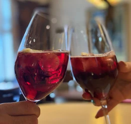 Zwei Personen stoßen mit Gläsern Rotwein in gemütlicher Umgebung an, leuchtender Farbton des Weins.