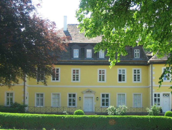 Historisches gelbes Herrenhaus mit weißen Fensterrahmen, umgeben von üppigem Grün und Bäumen.