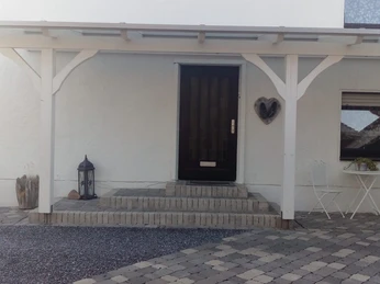 FeWo_Büsse_1.jpg Eingangsbereich eines Hauses mit überdachter Terrasse, hellem Pflaster und kleiner Sitzgruppe.