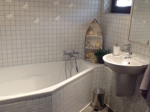 Badezimmer mit weiß gefliester Badewanne, Waschbecken, Spiegel und einem Regal mit Muschel-Deko.