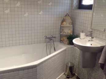 FeWo_Büsse_3.jpg Badezimmer mit weiß gefliester Badewanne, Waschbecken, Spiegel und einem Regal mit Muschel-Deko.