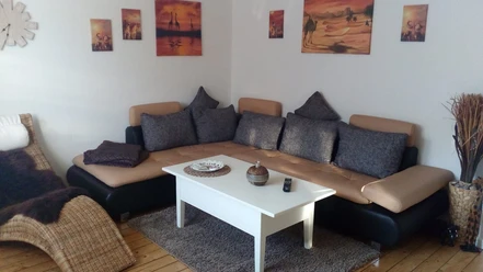 FeWo_Büsse_4.jpg Gemütliches Wohnzimmer mit beiger Couch, weißen Kaffeetisch und Wandbildern in warmen Tönen.