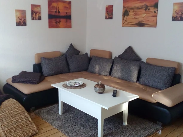 FeWo_Büsse_4.jpg Gemütliches Wohnzimmer mit beiger Couch, weißen Kaffeetisch und Wandbildern in warmen Tönen.