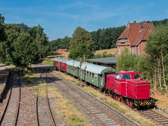 Bleckeder-kleinbahn-lok-foto_ccbysa_foto©heide-express.jpg Eine rote Lokomotive zieht historische, grüne und rote Waggons an einem sonnigen Tag entlang einer ländlichen Bahnstrecke. Im Hintergrund sind ein altes Backsteinhaus und grüne Bäume zu sehen, was der Szene einen nostalgischen, idyllischen Charakter verleiht.