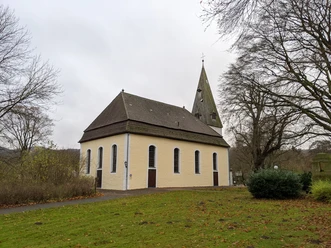 Kirche Stapelage_2©Tourist-Information Lage.jpg Die Kirche Stapelage mit spitzem Turm, umgeben von Bäumen und Rasen, unter bedecktem Himmel.