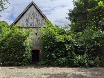 Steinbau mit steilem Dach, umgeben von üppigen Büschen und Bäumen unter bewölktem Himmel.