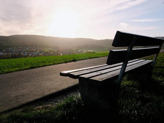 Aussichtspunkt Eine Holzbank steht am Wegesrand mit Blick über ein Dorf, beleuchtet von der Abendsonne.