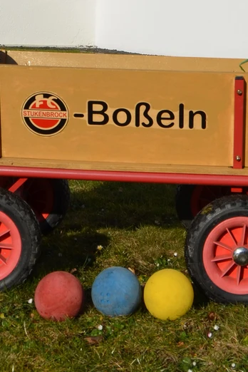 Ein Holzkarren auf Rasen mit Bällen in den Farben Rot, Blau und Gelb lässt auf ein Boßelspiel rückschließen.