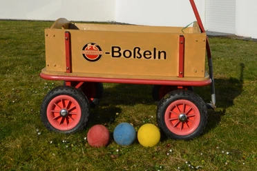 Boßeln_CC-BY-SA_Teutoburger_Wald_Stadt_Schloss_Holte-Stukenbrock.jpg Ein Holzkarren auf Rasen mit Bällen in den Farben Rot, Blau und Gelb lässt auf ein Boßelspiel rückschließen.