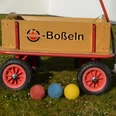 Boßeln_CC-BY-SA_Teutoburger_Wald_Stadt_Schloss_Holte-Stukenbrock.jpg Ein Holzkarren auf Rasen mit Bällen in den Farben Rot, Blau und Gelb lässt auf ein Boßelspiel rückschließen.