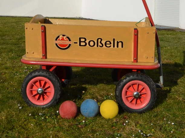 Boßeln_CC-BY-SA_Teutoburger_Wald_Stadt_Schloss_Holte-Stukenbrock.jpg Ein Holzkarren auf Rasen mit Bällen in den Farben Rot, Blau und Gelb lässt auf ein Boßelspiel rückschließen.