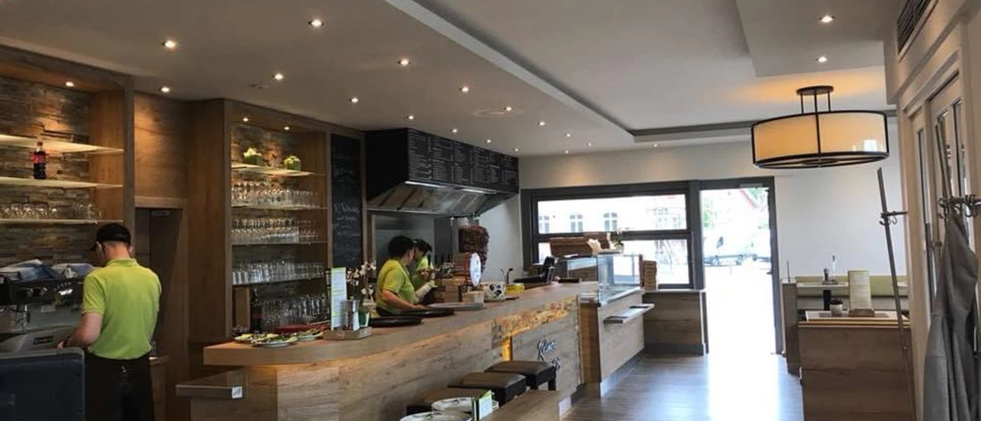 Modernes Café mit Holztresen, großen Fenstern und zwei Beschäftigten in grünen Hemden.