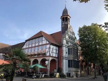 Rathaus Nienburg mit Fachwerk und Uhrturm, umgeben von Bäumen und einem Café im Vordergrund.Nienburg town hall with half-timbered building and clock tower, surrounded by trees and a café in the foreground.Nienburgs rådhus med bindingsværksbygning og klokketårn, omgivet af træer og en café i forgrunden.Stadhuis van Nienburg met vakwerkgebouw en klokkentoren, omringd door bomen en een café op de voorgrond.