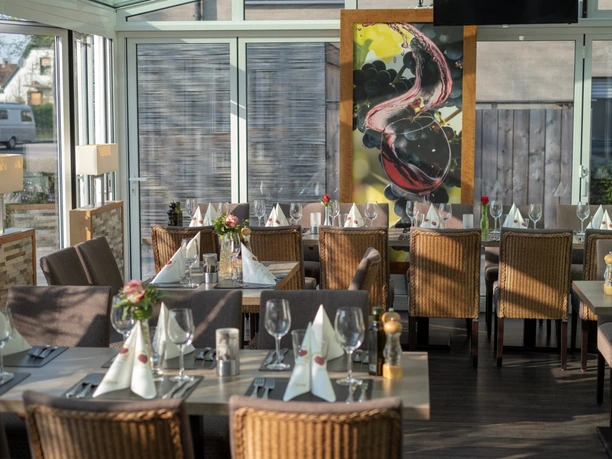 Mykonos_3.png Ein lichtdurchfluteter Gastraum mit eingedeckten Tischen, Rattan-Stühlen und großem Gemälde an der Wand.