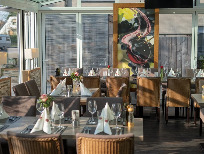 Mykonos_3.png Ein lichtdurchfluteter Gastraum mit eingedeckten Tischen, Rattan-Stühlen und großem Gemälde an der Wand.