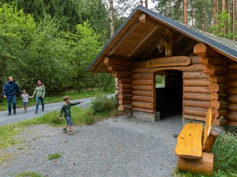Drachenrundweg Holzhütte am Waldrand, Familie mit zwei Kindern spaziert auf einem Kiesweg vorbei.Wooden hut at the edge of the forest, family with two children walking past on a gravel path.Dřevěná chata na okraji lesa, kolem jde po štěrkové cestě rodina se dvěma dětmi.Drewniana chata na skraju lasu, rodzina z dwójką dzieci przechodząca obok żwirową ścieżką.Houten hut aan de rand van het bos, familie met twee kinderen lopen voorbij over een grindpad.Capanna di legno ai margini del bosco, famiglia con due bambini che passa su un sentiero di ghiaia.