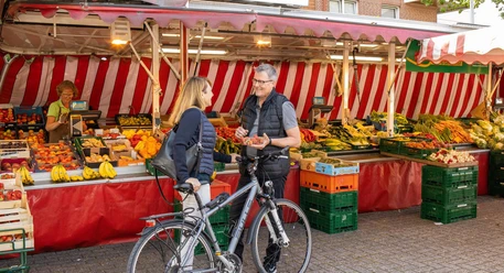 wochenmarkt-bad-zwischenahn-beusch-mit-fahrrad.jpg Eine Frau und ein Mann unterhalten sich auf einem Wochenmarkt in Bad Zwischenahn. Im Hintergrund sind zahlreiche Obst- und Gemüsestände zu sehen. Der Mann hält eine Schale mit Erdbeeren, während beide eine entspannte Atmosphäre genießen.
