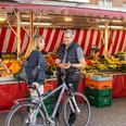wochenmarkt-bad-zwischenahn-beusch-mit-fahrrad.jpg Eine Frau und ein Mann unterhalten sich auf einem Wochenmarkt in Bad Zwischenahn. Im Hintergrund sind zahlreiche Obst- und Gemüsestände zu sehen. Der Mann hält eine Schale mit Erdbeeren, während beide eine entspannte Atmosphäre genießen.