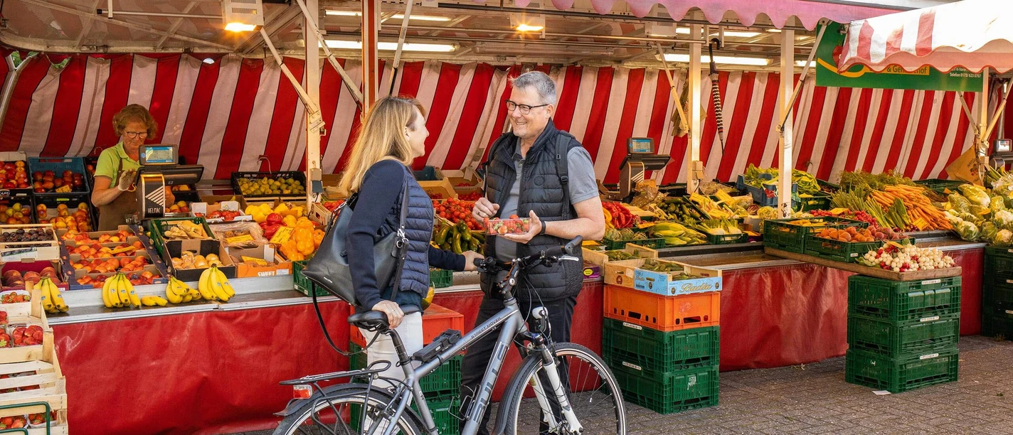 wochenmarkt-bad-zwischenahn-beusch-mit-fahrrad.jpg A woman and a man are chatting at a weekly market in Bad Zwischenahn. Numerous fruit and vegetable stalls can be seen in the background. The man is holding a bowl of strawberries while they both enjoy the relaxed atmosphere.