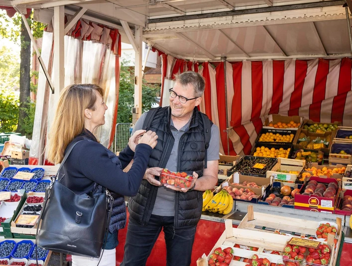 wochenmarkt-bad-zwischenahn-obst.jpg Ein Wochenmarktstand mit frischem Obst: Verkäufer reicht einer Kundin lächelnd Erdbeeren.