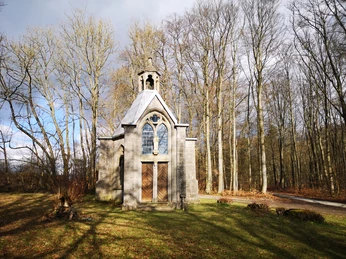 Kleine Steinkapelle mit Glockenturm, umgeben von Bäumen, steht im lichten Schatten eines Waldes.